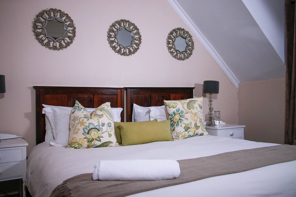 Cama bem arrumada com almofadas florais, luminária de cabeceira e três espelhos na parede.