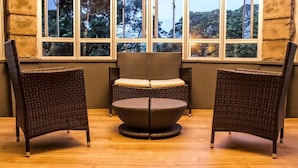 A room with a wicker chair and a round table with a cushion, positioned in front of a window with a view of trees.