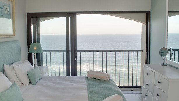 Un dormitorio con cama, almohadas, una mesita de noche, lámpara, cómoda y un ventanal con vista al océano.