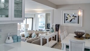 Una cocina moderna con armarios blancos, un comedor con mobiliario blanco y una sala de estar con sofá color beige.