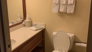 A bathroom with a toilet, sink, and mirror. There are towels hanging on the wall and a trash can.