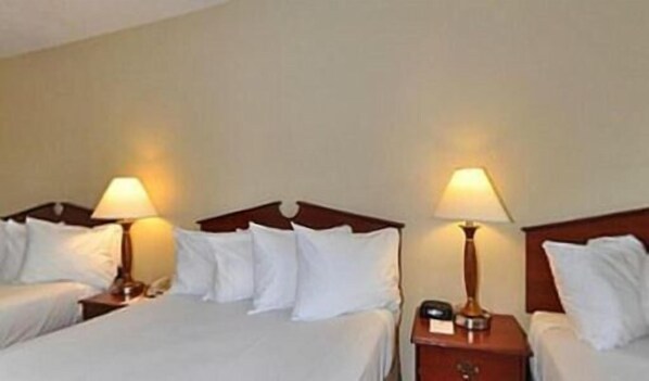 Habitación de hotel con dos camas, cada una con sábanas blancas y cabeceros de madera. Hay dos mesitas de noche, una de ellas con una lámpara y un reloj pequeño.