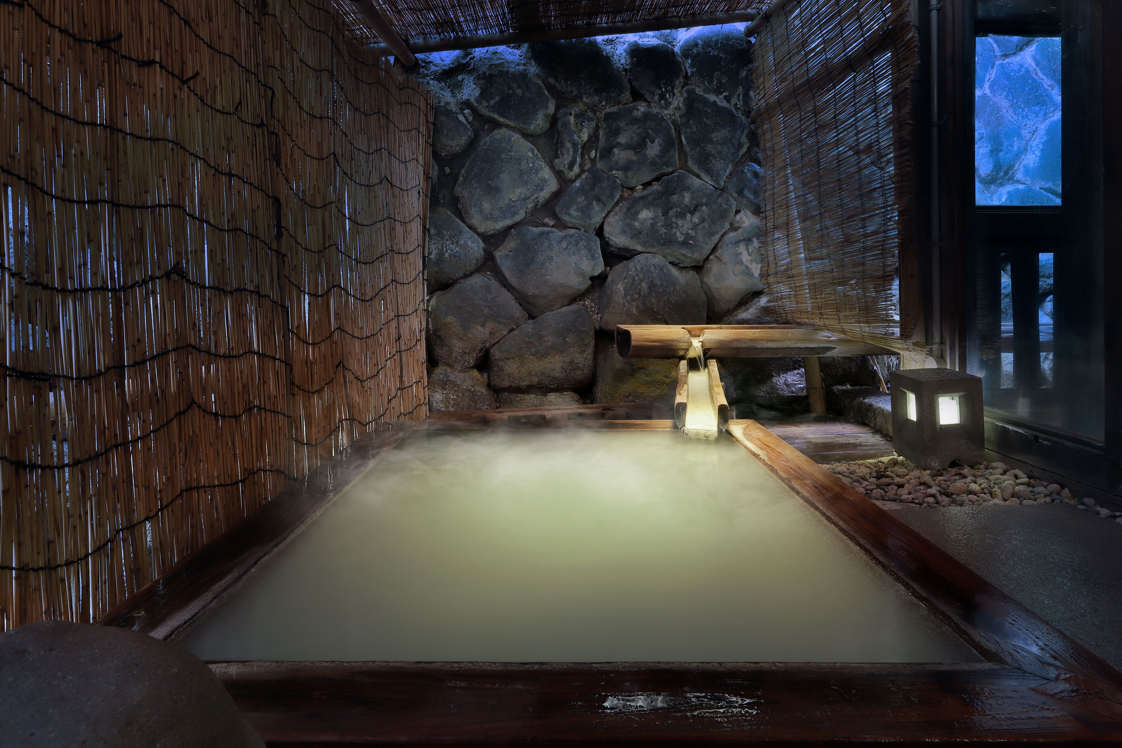 Un onsen japonés tradicional con pared de piedra, banco de madera y cuenca de piedra.