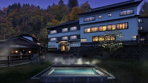 A modern hotel with multiple floors, a swimming pool, and a view of a forested hillside. - Omiya Ryokan (Yamagata)