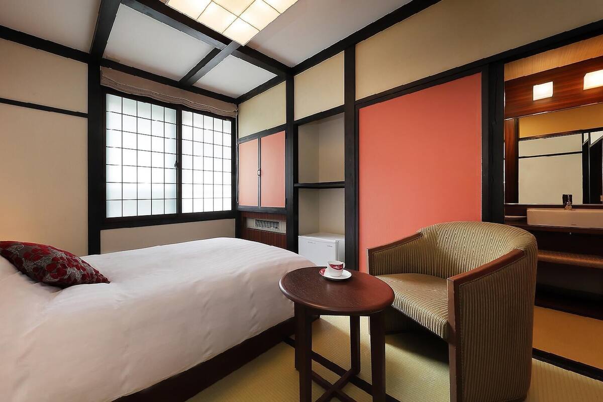 A hotel room with a bed, a chair, a small table, a window, and a mirror.