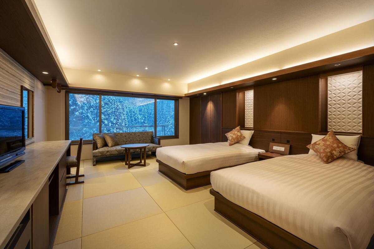 Una habitación de hotel moderna con dos camas, un sofá, un televisor y ventanas amplias con vistas nevadas.