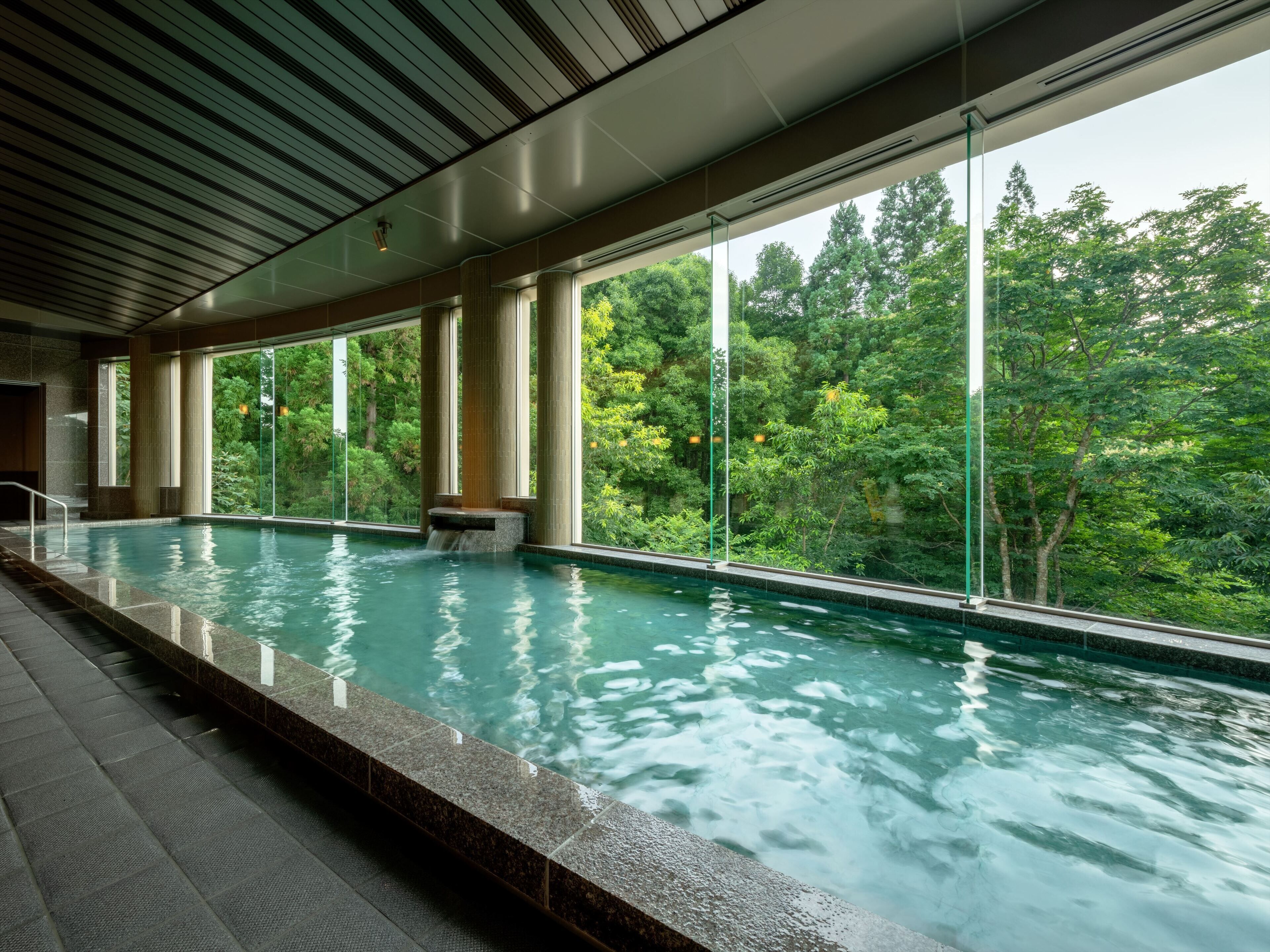 An indoor swimming pool with clear glass walls, large windows, and a view of lush greenery outside.