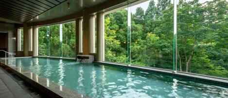 An indoor swimming pool with clear glass walls, large windows, and a view of lush greenery outside.