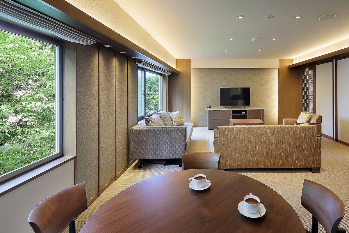 A modern hotel room with a sofa, a dining table with two cups of coffee, a TV, and a view of greenery outside.