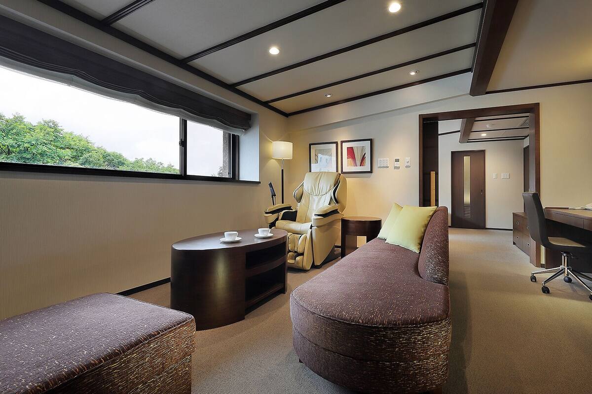A hotel room with a massage chair, a desk, a sofa, and a small table with cups.