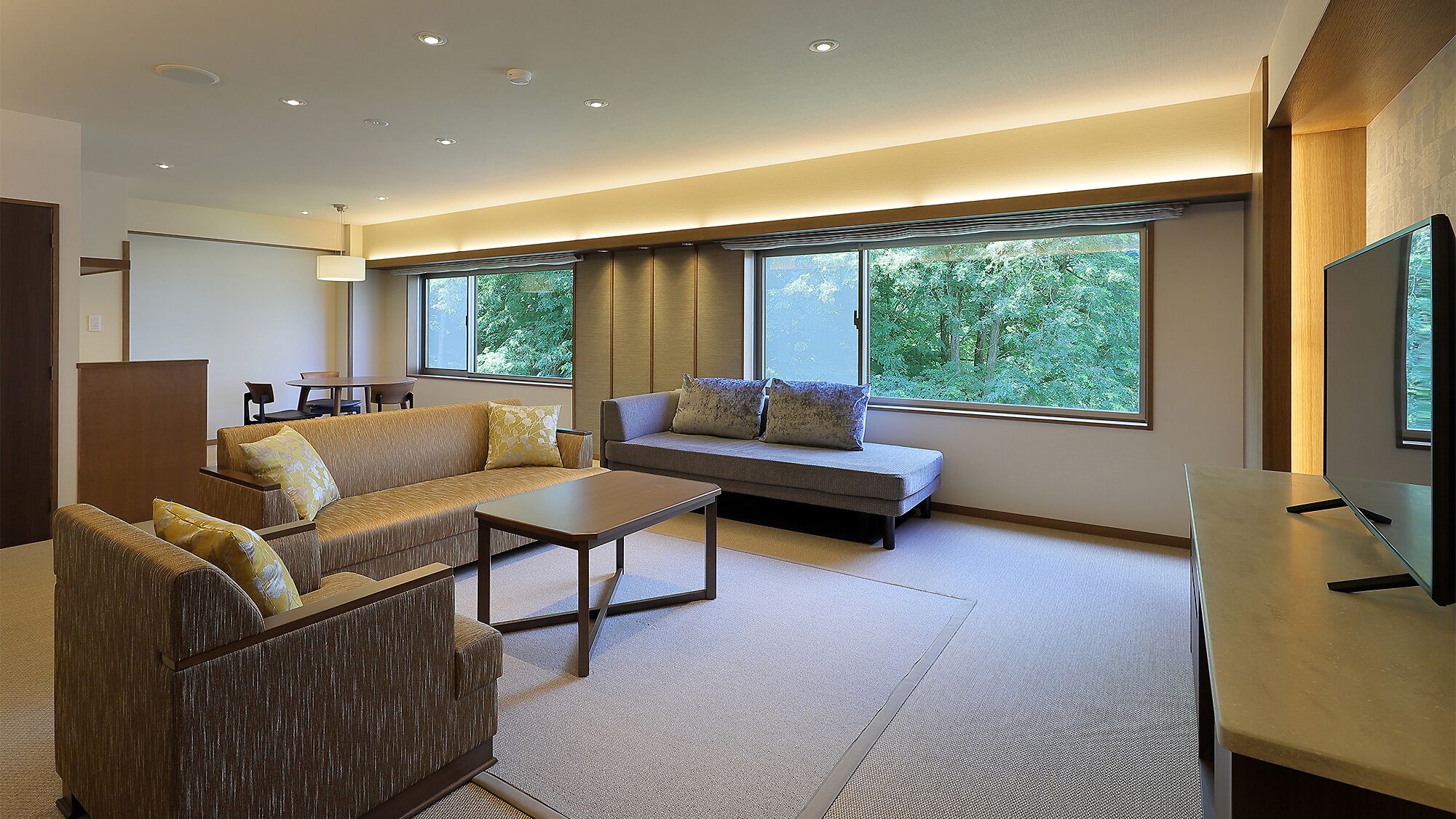 A modern living room with a sofa, armchair, and coffee table. There are large windows with a view of trees.