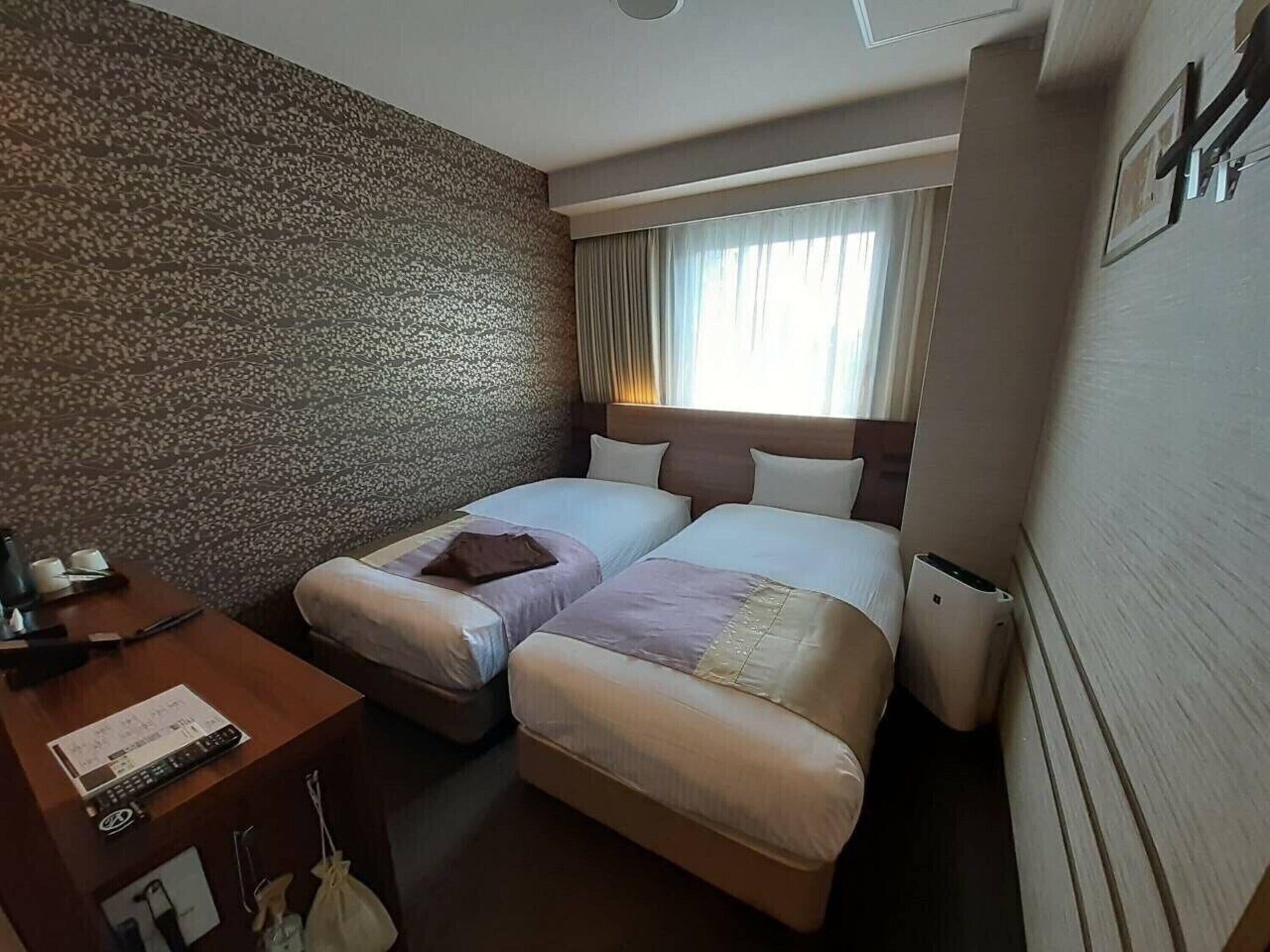 A hotel room with two beds, a desk with a telephone and a notepad, a window with curtains, and a wall with a textured pattern.