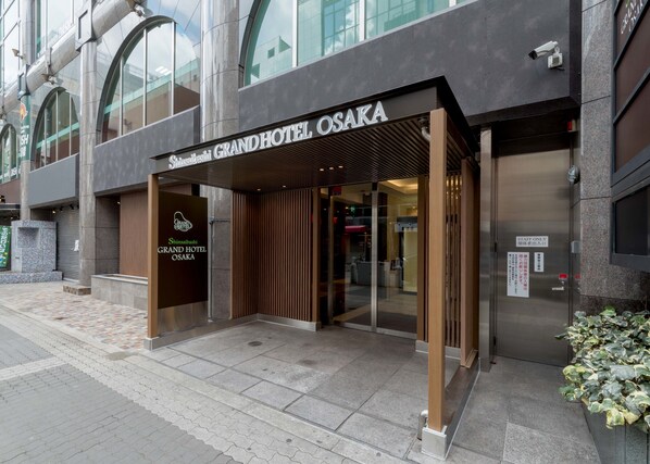 The exterior of the Shinsaibashi Grand Hotel Osaka with a modern facade, a wooden entrance canopy, and a visible elevator.