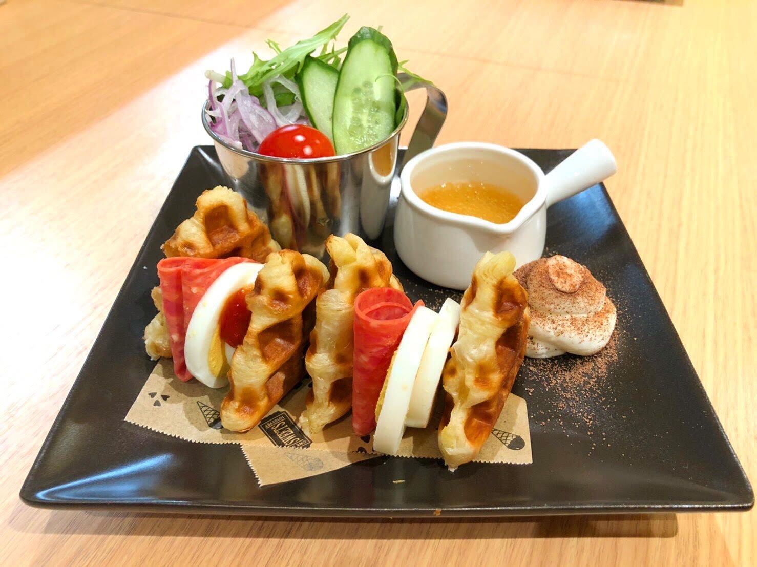 A square black plate with waffles, a small salad, a cup of sauce, and a piece of bread.