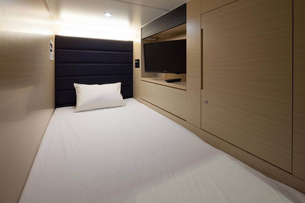 A neatly made bed with a white pillow and a dark headboard in a modern room with wooden walls and a built-in TV.