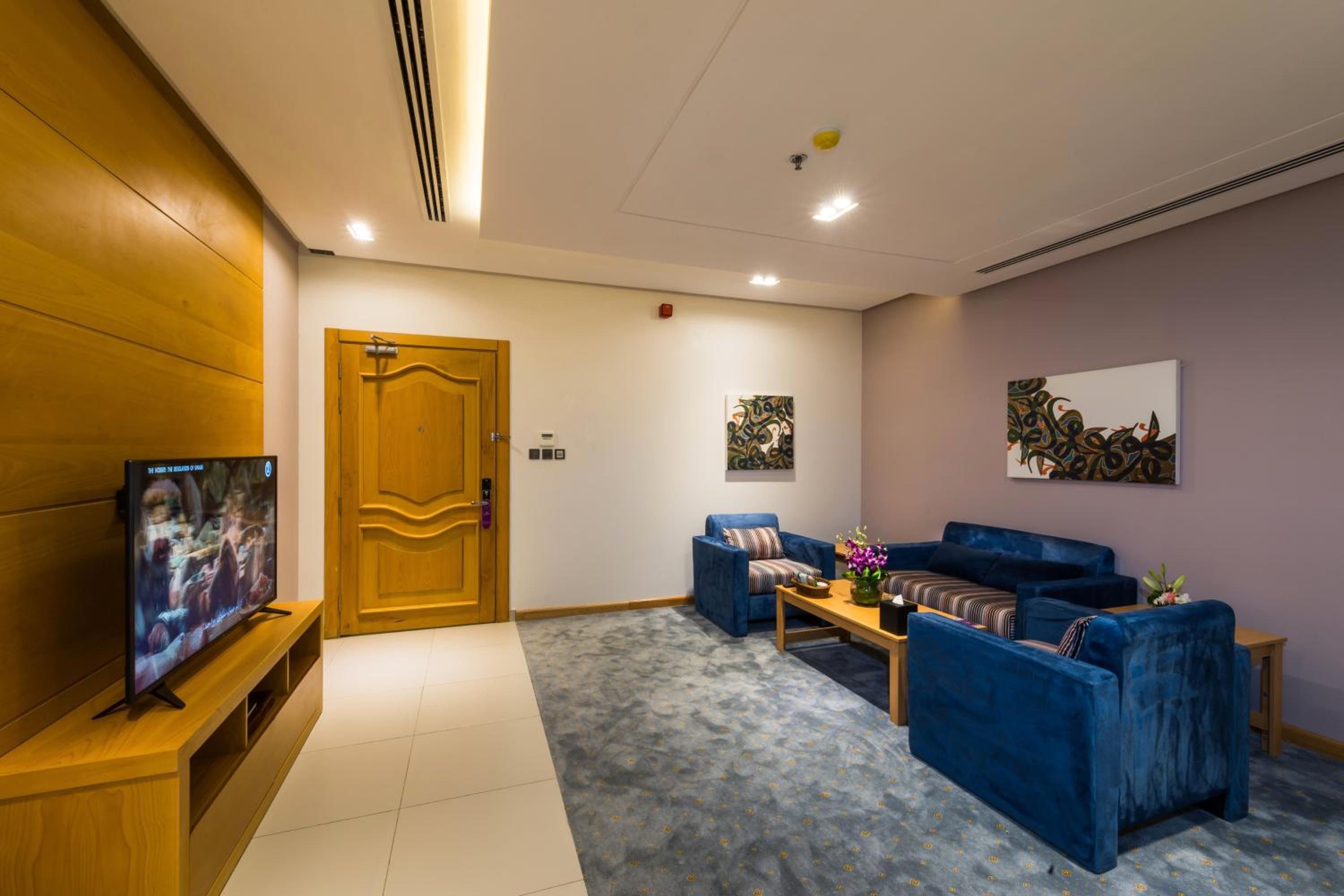 A hotel room with a television on a wooden stand, a blue sofa set, a small table with a vase of flowers, and two wall-mounted paintings.