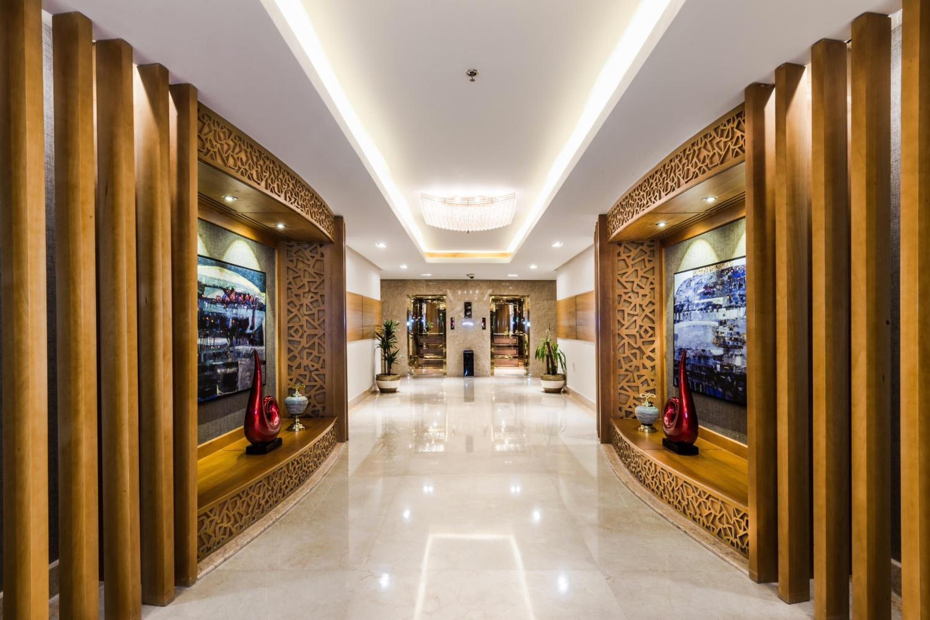A spacious, well-lit hallway with a polished floor, flanked by wooden panels with intricate designs, leading to a reception area with a chandelier and a large framed artwork on the wall.