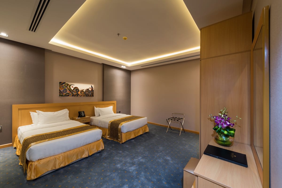 A hotel room with two beds, a desk, a chair, a vase with flowers, and a painting on the wall.