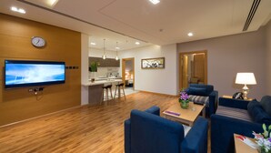 A modern hotel room with a wooden floor, a flat-screen TV mounted on the wall, a dining area with bar stools, a sofa, and a lamp. - Boudl Al Munsiyah (Riyadh)
