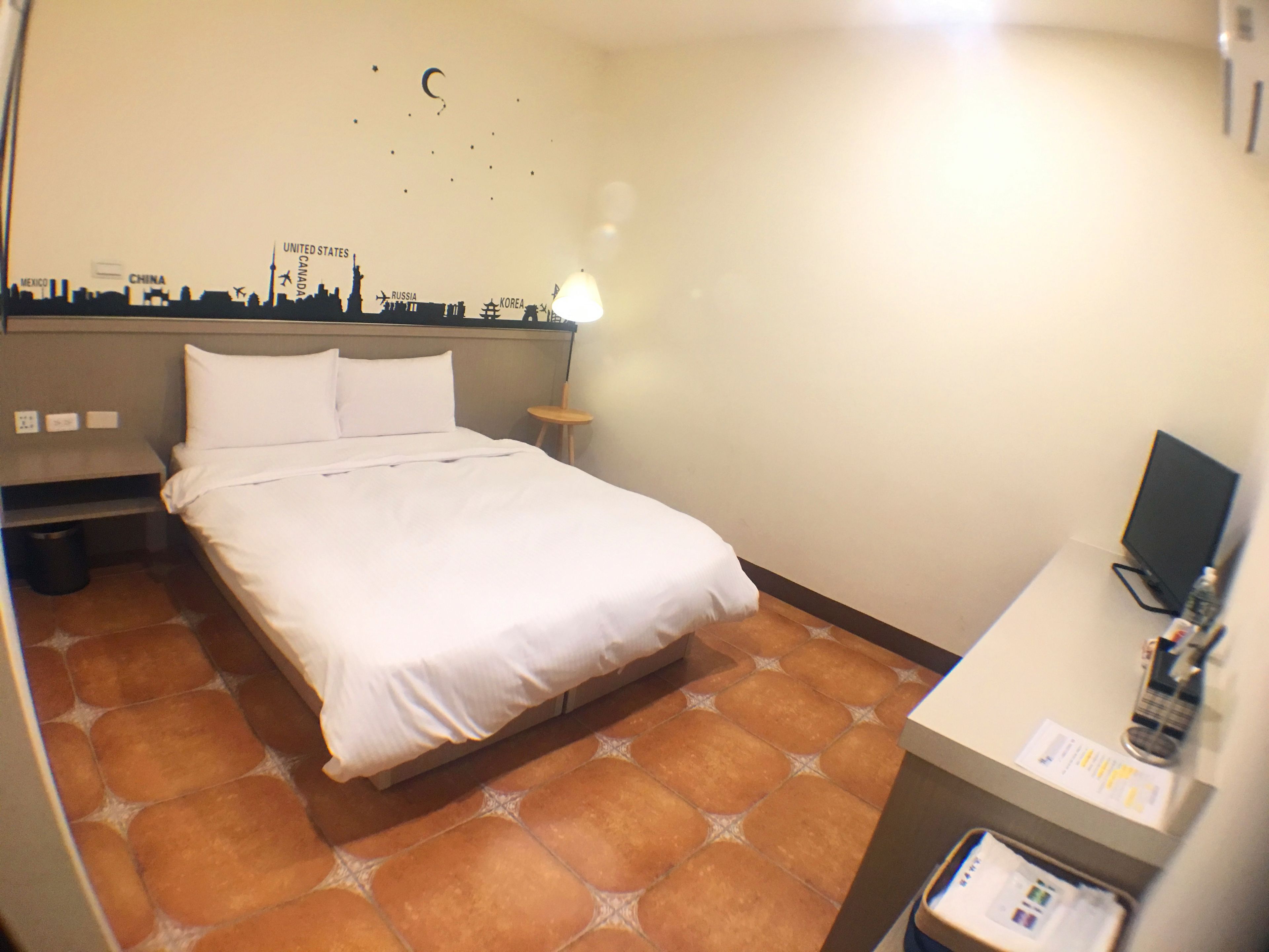 A hotel room with a bed, a nightstand, a small table, and a wall with a city skyline mural.