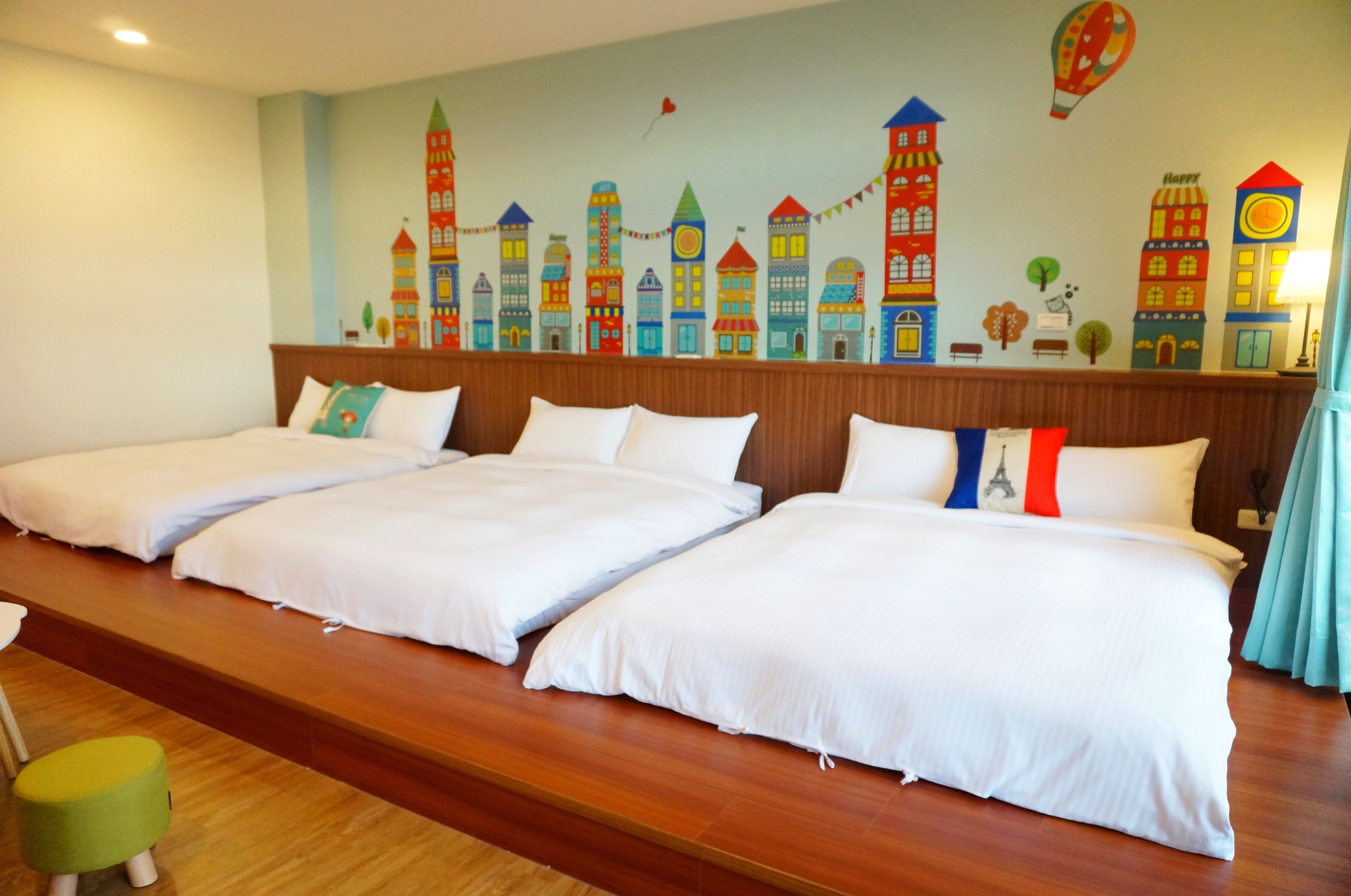 Een hotelkamer met drie bedden, een houten hoofdeinde, een kleurrijke muurschildering van een stadsgezicht en een klein, groen voetenbankje.