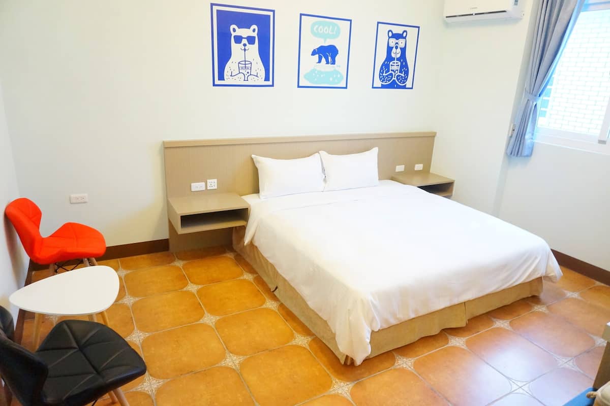 A hotel room with a bed, a nightstand, a chair, and a table. There are three framed pictures on the wall.