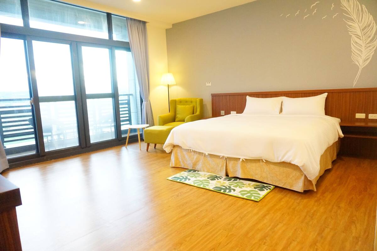 A hotel room with a large bed, a yellow armchair, a floor lamp, and a view through large windows.