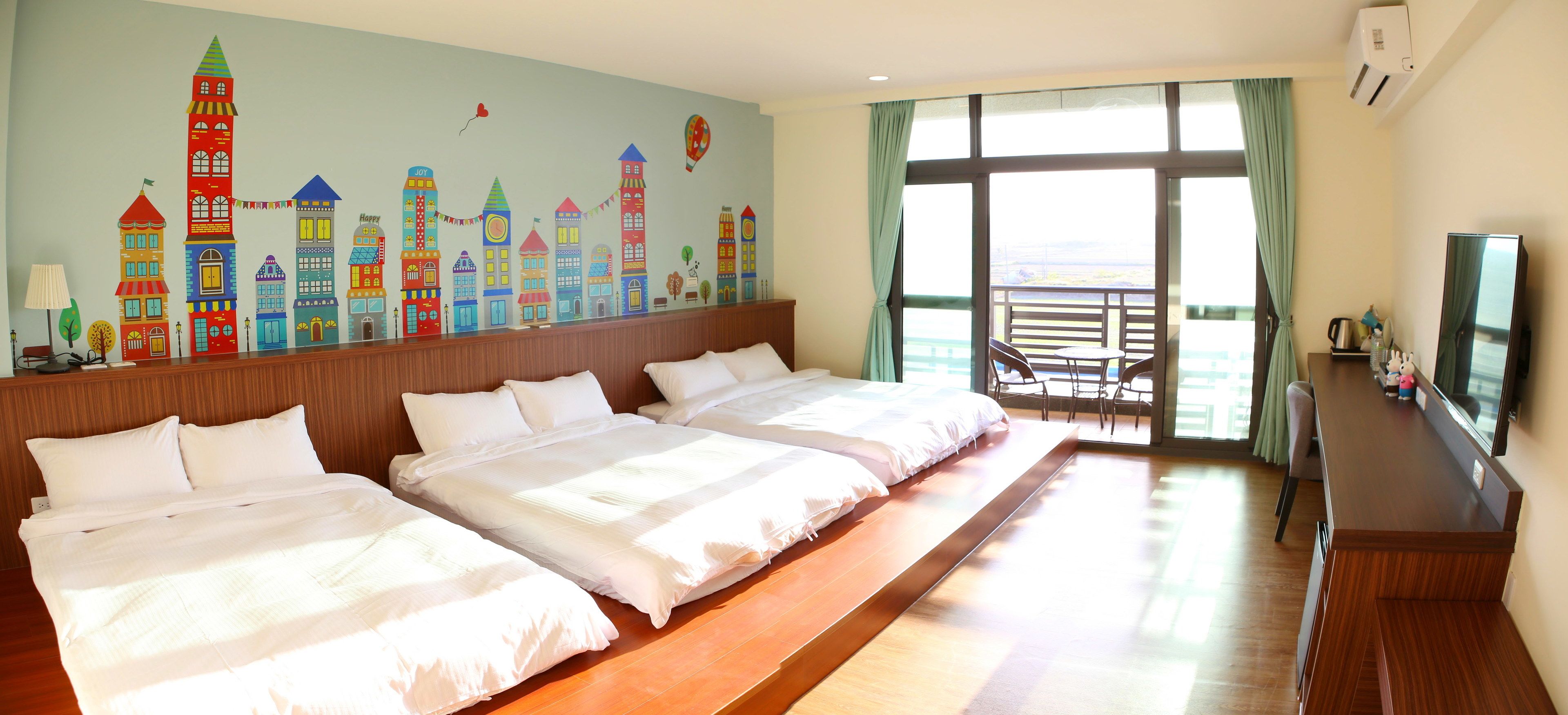 A hotel room with two beds, a TV, a balcony with a view, and a colorful cityscape mural on the wall.