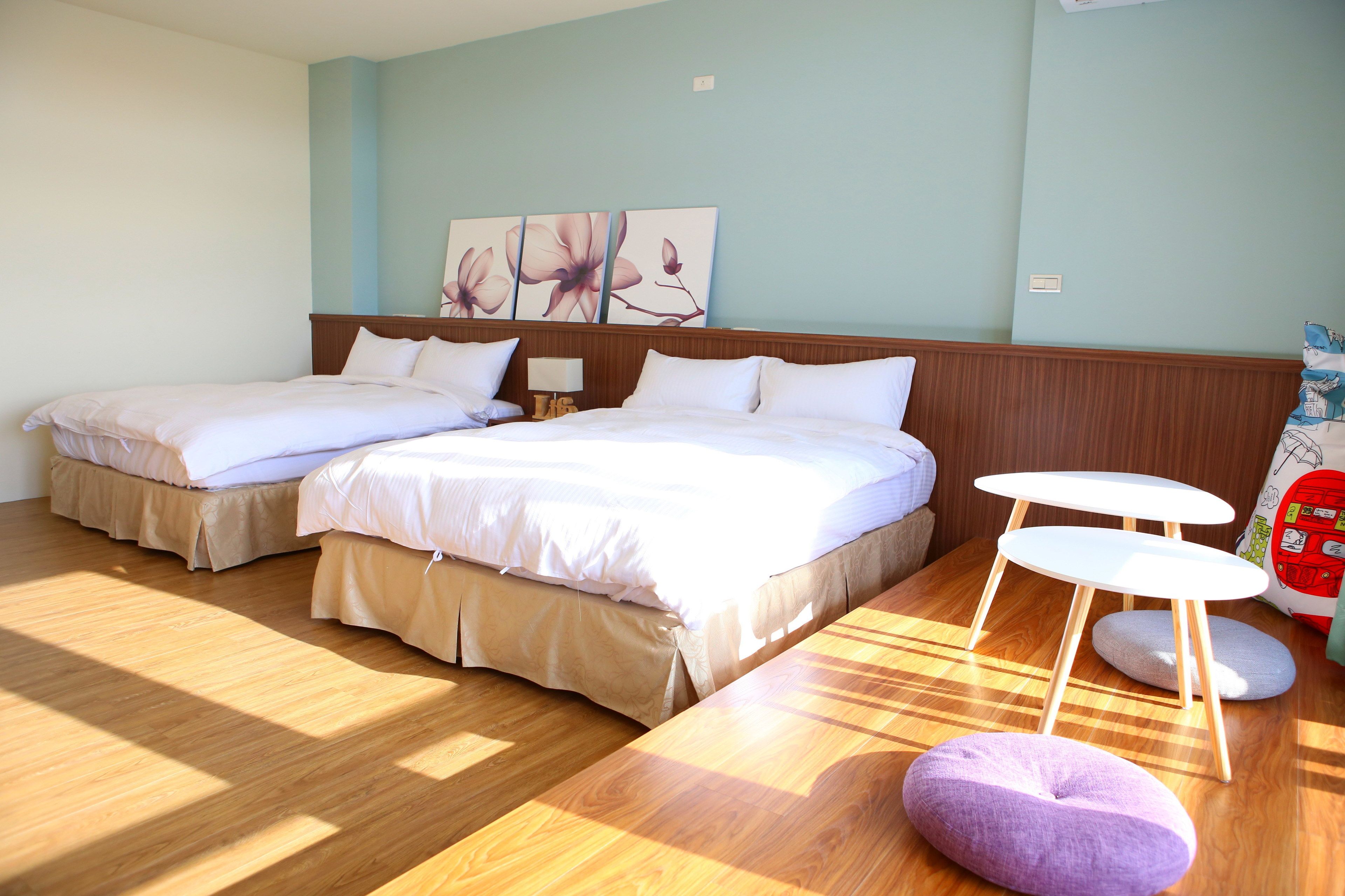 A hotel room with two beds, a wooden floor, a small table, and a wall with floral artwork.