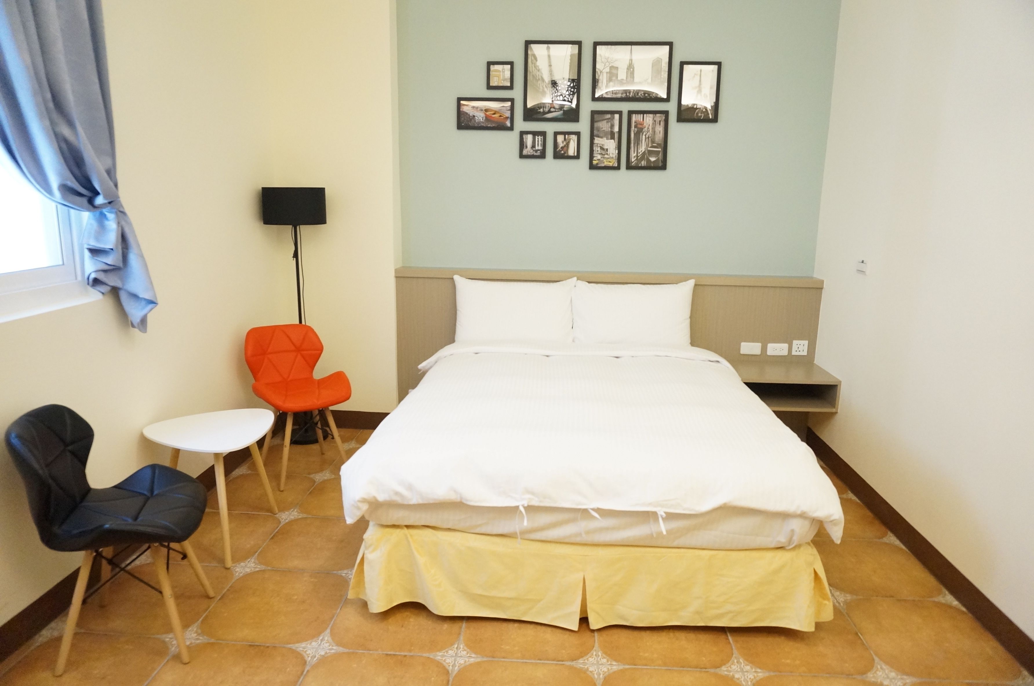 A hotel room with a bed, a chair, a small table, and a wall with framed pictures.