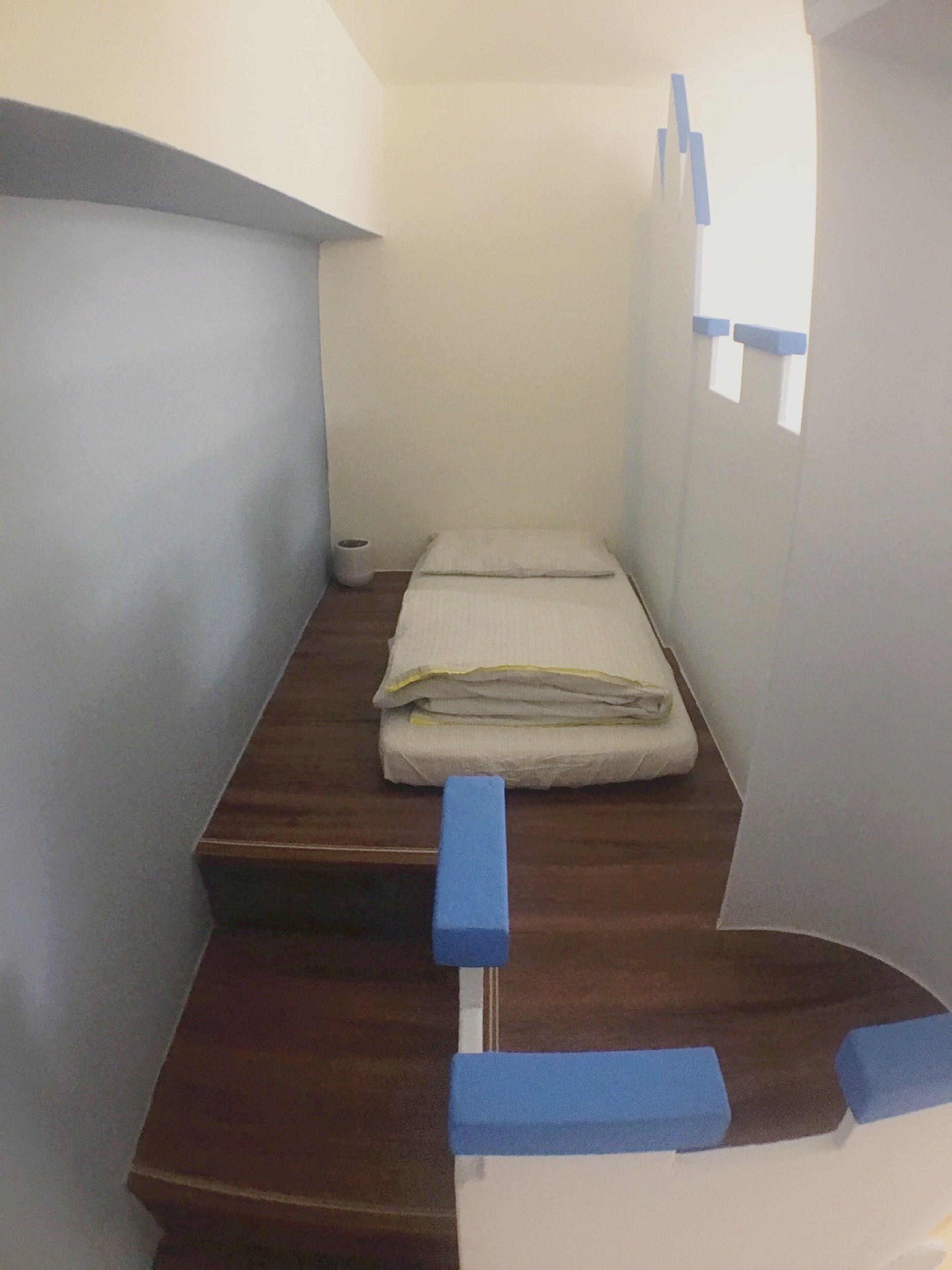 Una habitación pequeña y estrecha con una cama individual, un banco de madera y un ventanal.