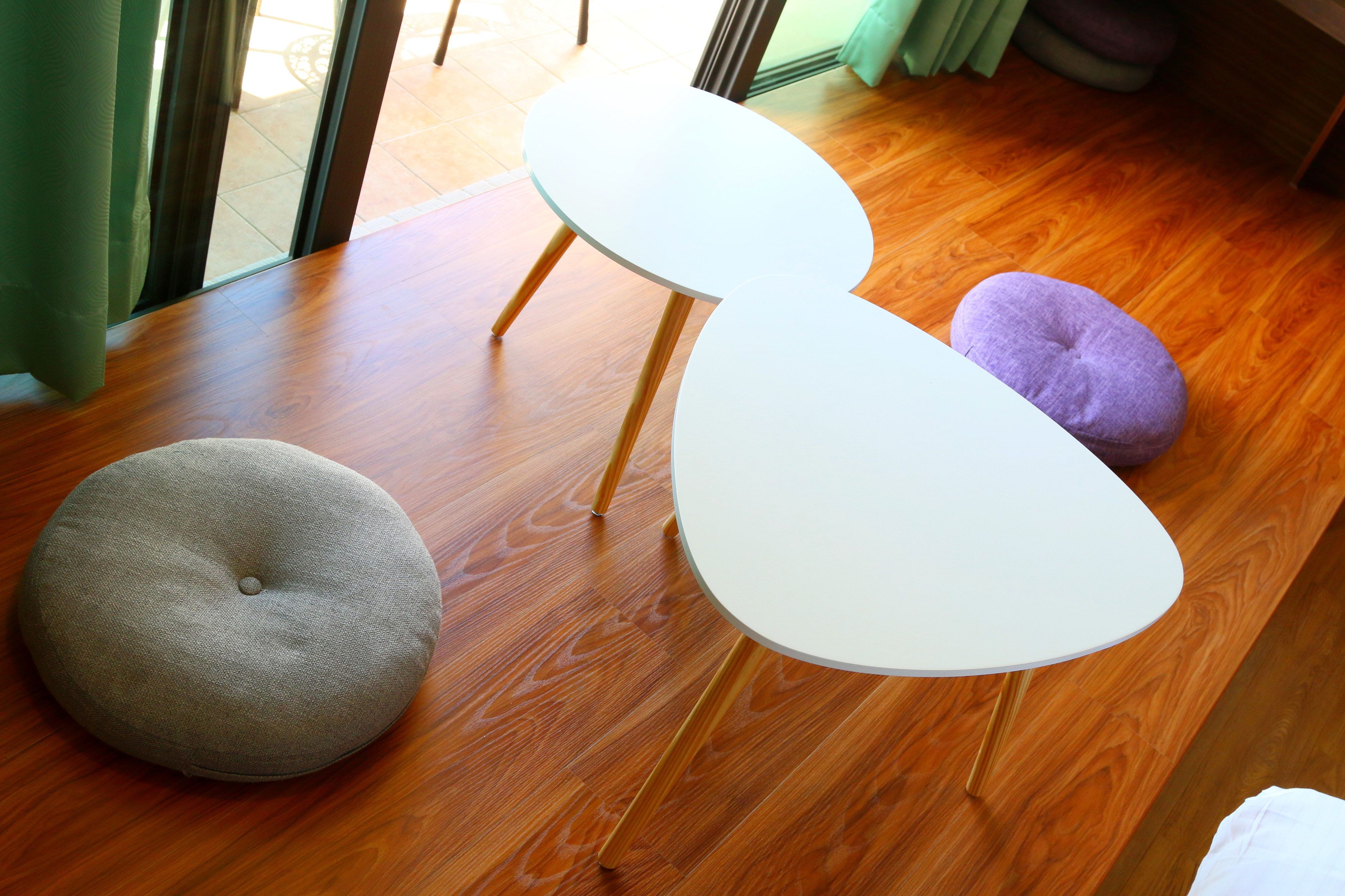 A room with a wooden floor, a white table with wooden legs, and two round ottomans, one grey and one purple.
