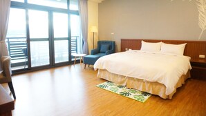 Habitación de hotel con una cama grande, un sillón azul, una mesita pequeña y vistas al mar a través de amplias ventanas.