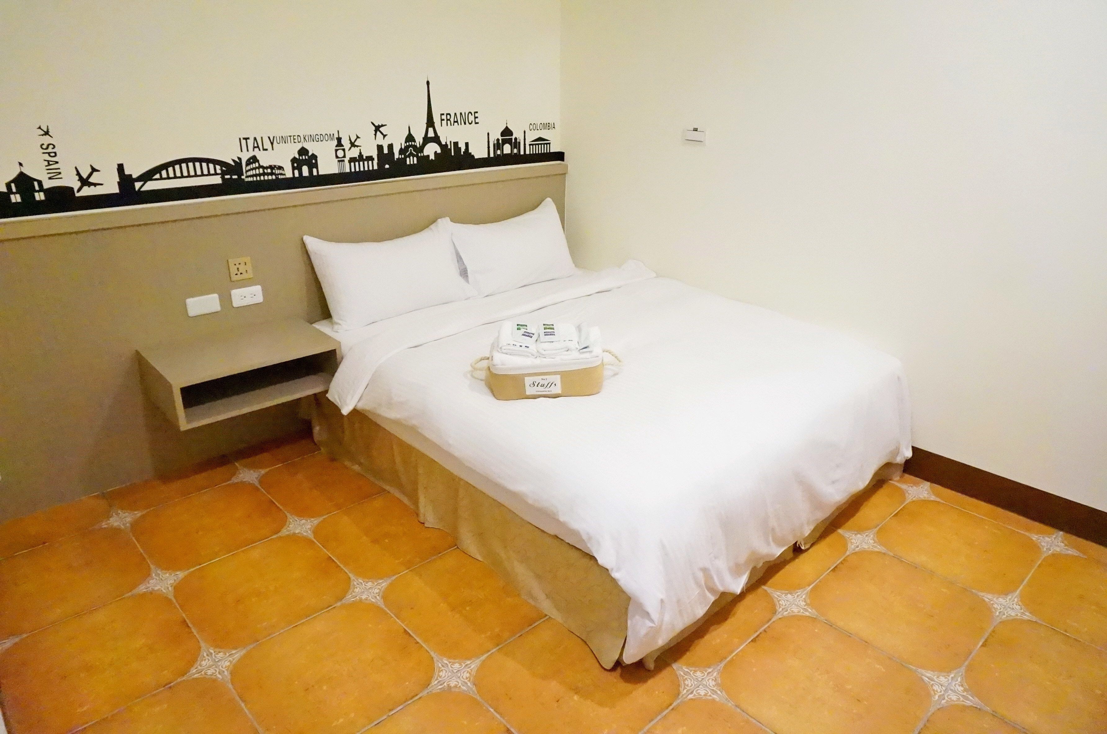 A hotel room with a bed, a small bedside table, a wall decoration with city names, and a tiled floor.