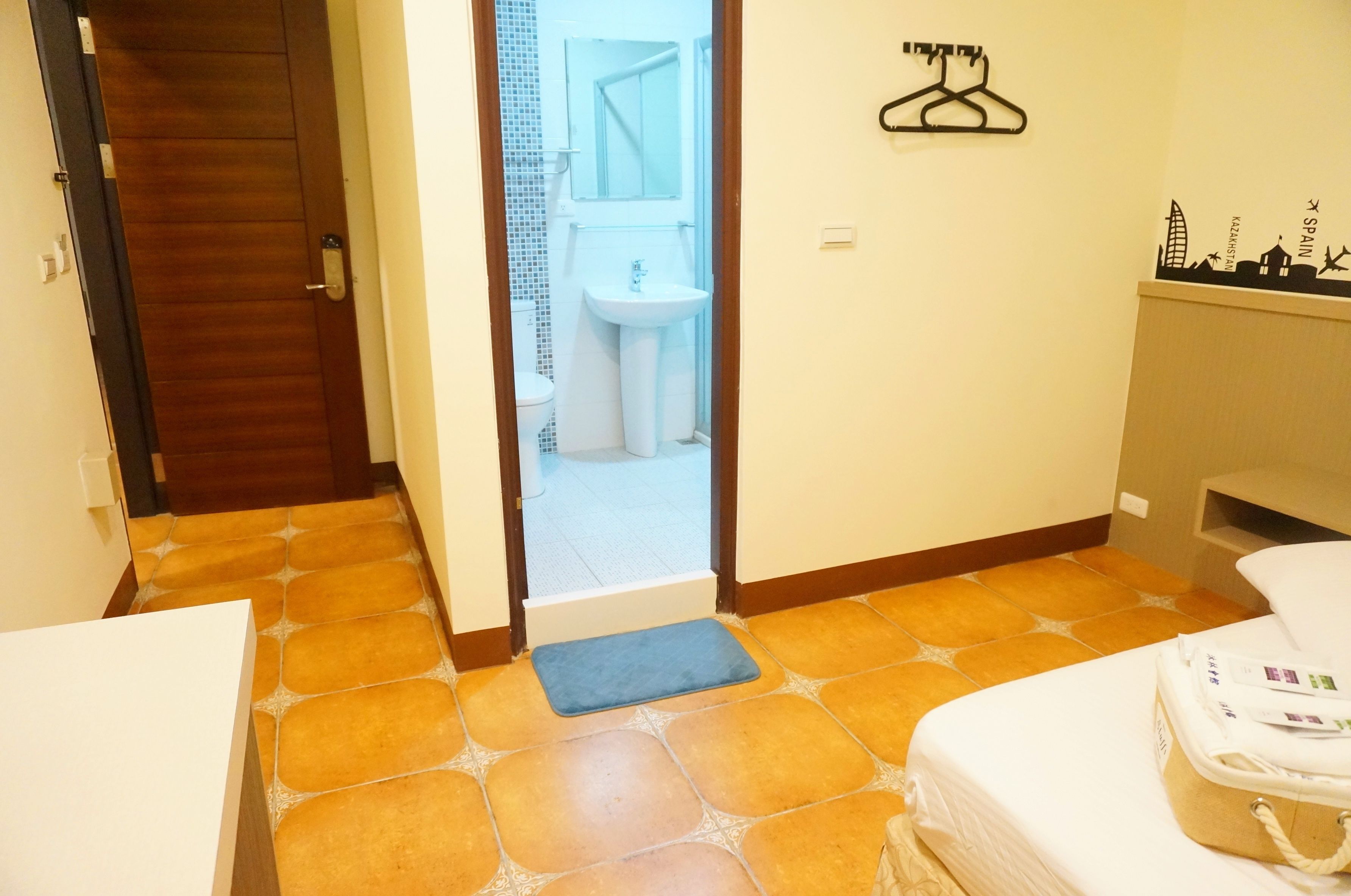 A hotel room with a wooden door, a bathroom visible through an open door, a bed, a bedside table, and a wall-mounted towel rack.