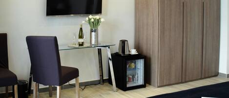 A modern hotel room with a flat-screen TV mounted on the wall, a glass desk with a chair, a minibar, and a wardrobe.