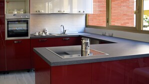 A modern kitchen with red cabinets, a built-in oven, a stainless steel sink, and a window with a view of a brick wall. - Gregori Guest House (Rome)