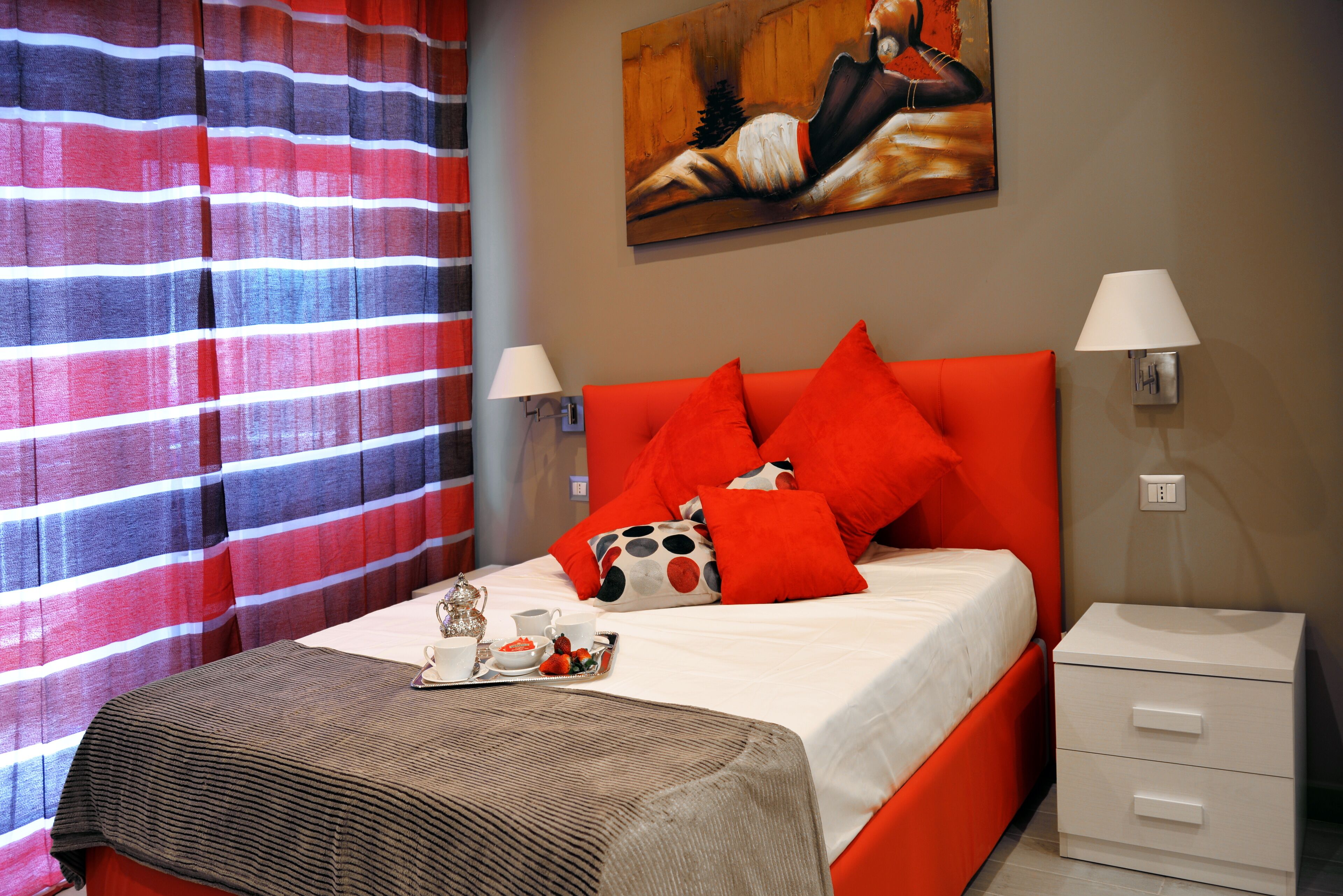 Camera d'albergo con un letto, un comodino, una lampada, un quadro appeso al muro e una finestra con tende a righe.