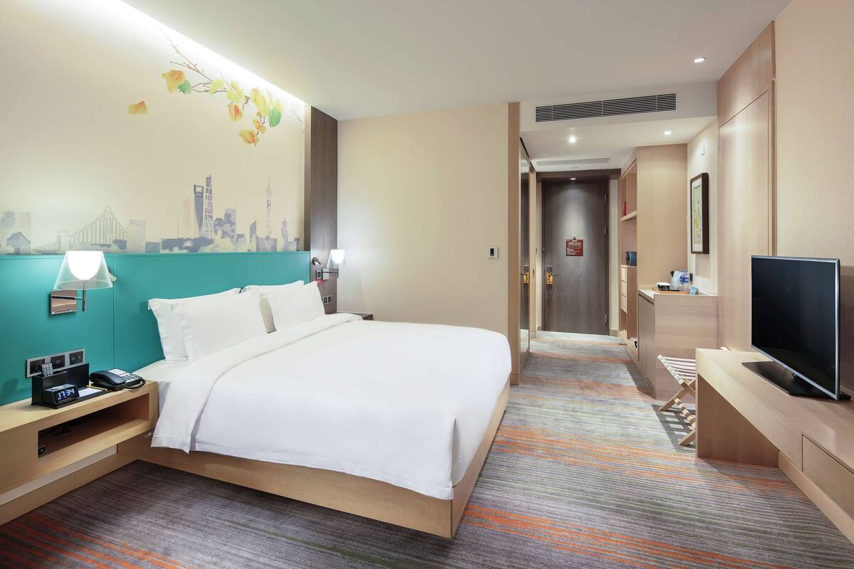 A hotel room with a bed, a desk with a lamp, a TV, and a cityscape mural on the wall.