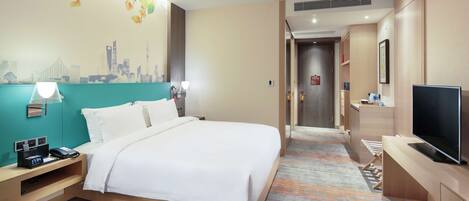A hotel room with a bed, a desk with a lamp, a TV, and a cityscape mural on the wall.