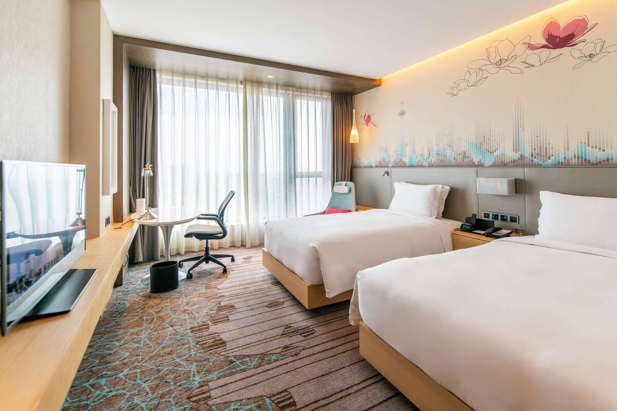 A hotel room with two beds, a desk, a chair, a TV, and a wall with a floral mural.