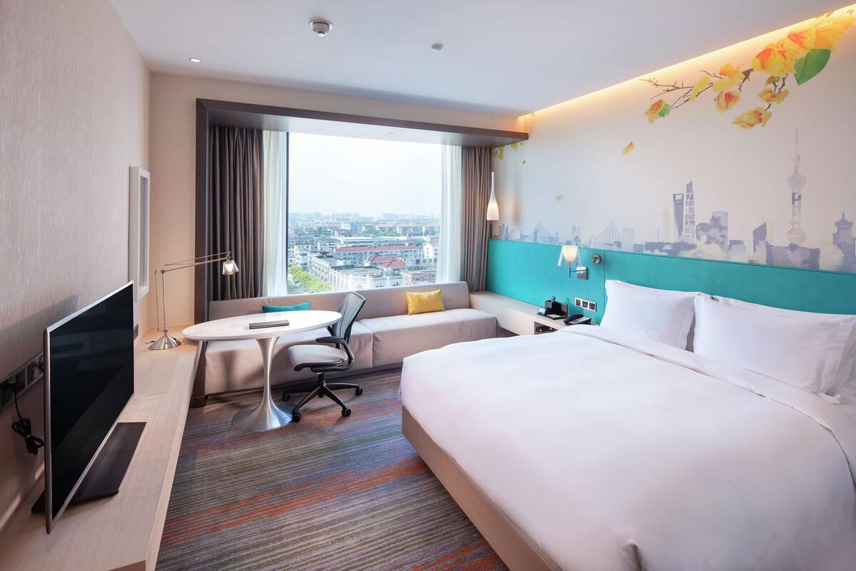 A hotel room with a large bed, a desk with a chair, a TV, and a view of the city through the window.