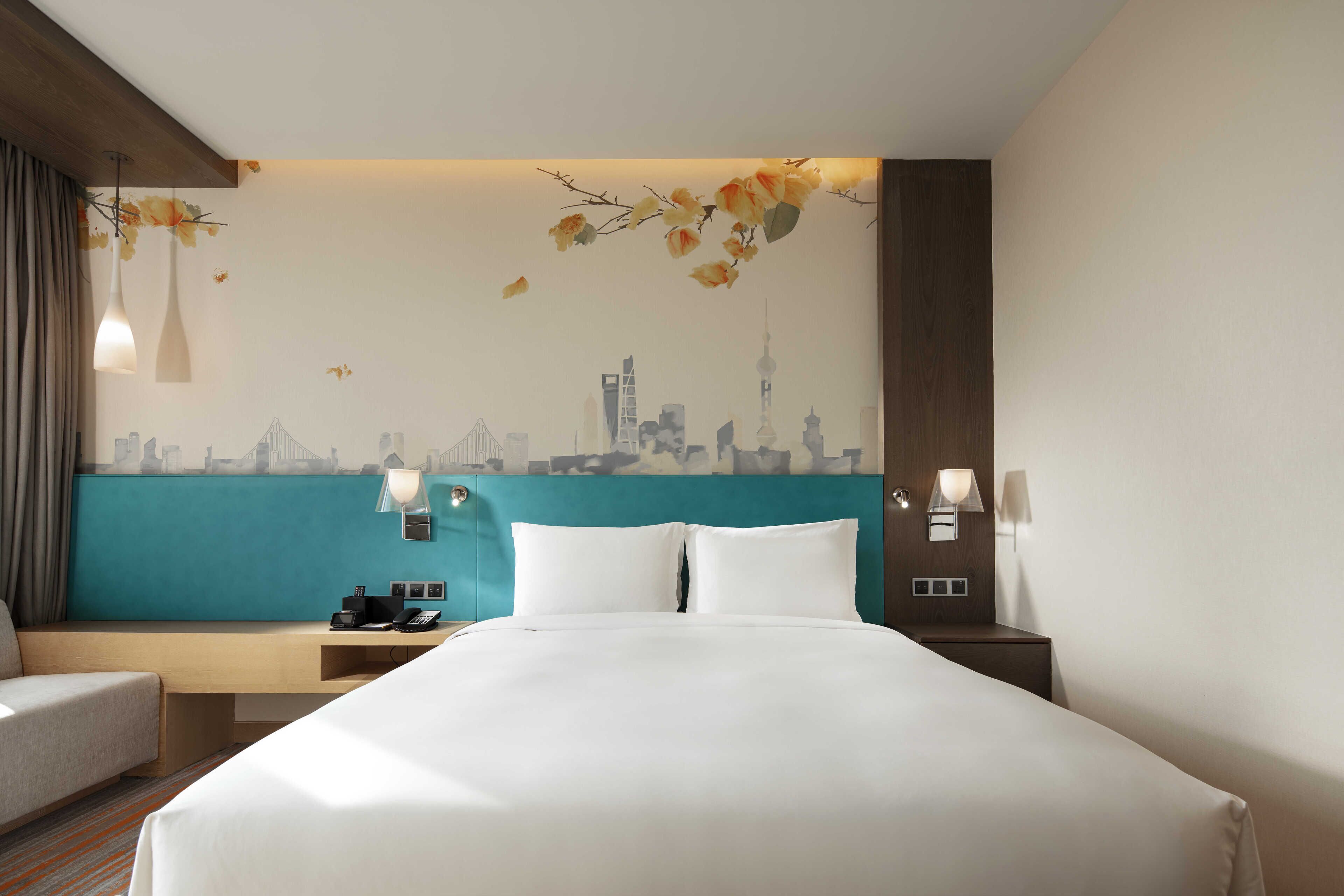 A hotel room with a bed, a desk, a chair, a nightstand, and a wall with a cityscape mural.