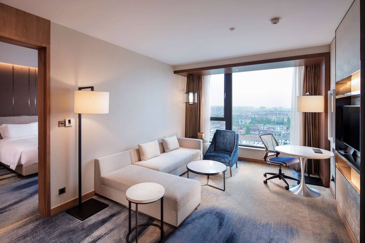 A hotel room with a sofa, two side tables, a chair, a TV, and a large window with a city view.