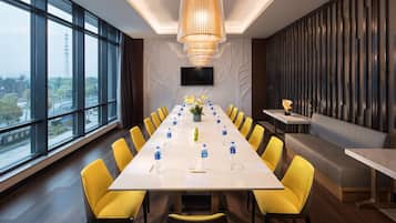 Sala de conferências com uma mesa comprida, cadeiras amarelas, um lustre moderno, uma TV na parede e vista para a cidade.