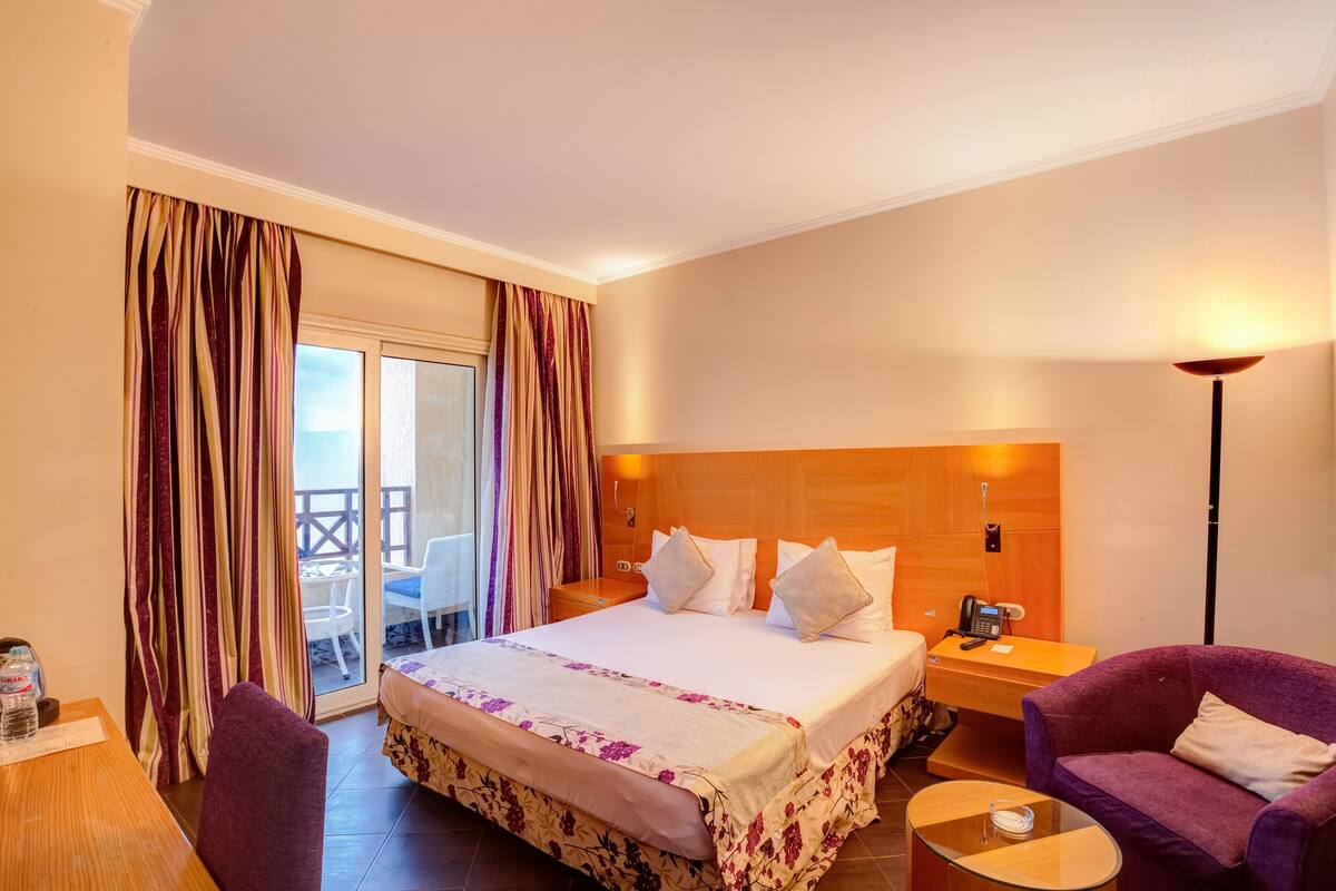 Habitación de hotel con cama, escritorio, silla, sofá púrpura y balcón con cortinas.