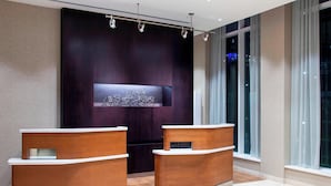 A reception area with two wooden reception desks, a dark wall with a geometric art piece, and large windows with city lights visible outside. - Courtyard by Marriott New York Queens/Fresh Meadows (Fresh Meadows)