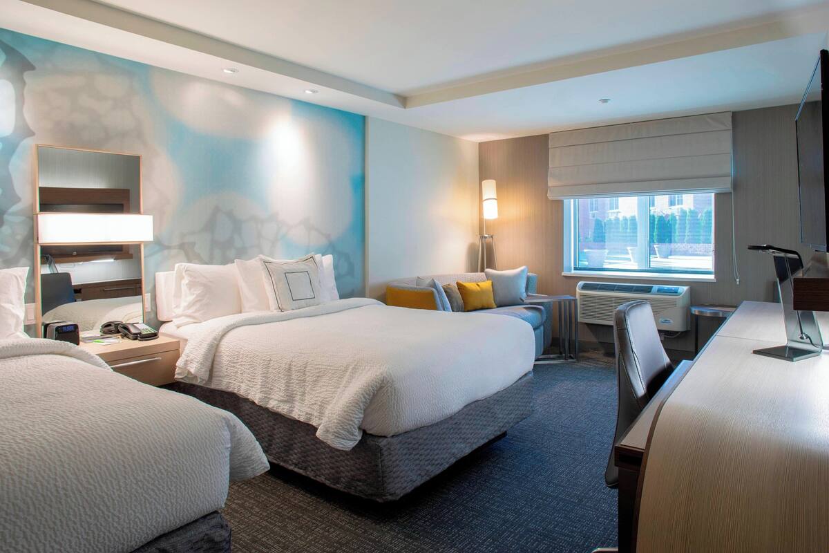 A hotel room with two beds, a desk, a chair, a lamp, a window with a view, and an air conditioning unit.