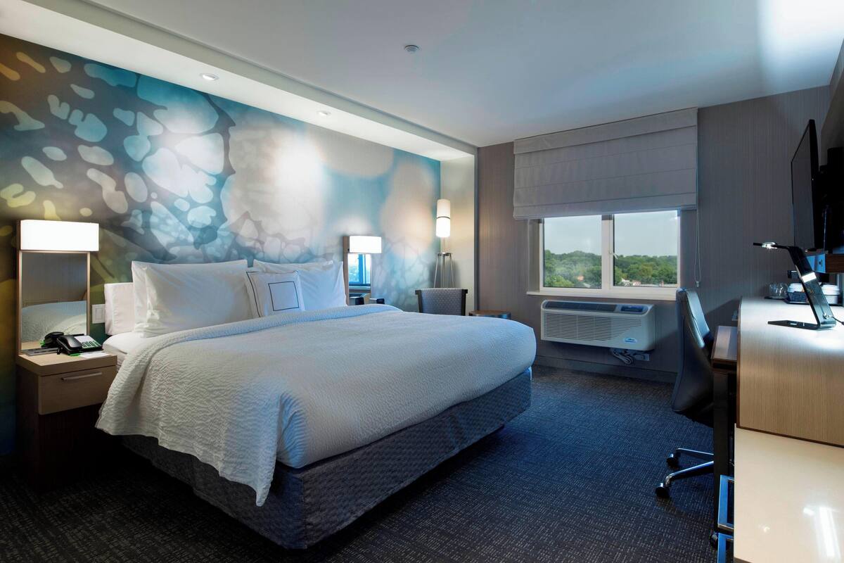 A hotel room with a large bed, bedside tables with lamps, a desk with a computer, and a window with a view.
