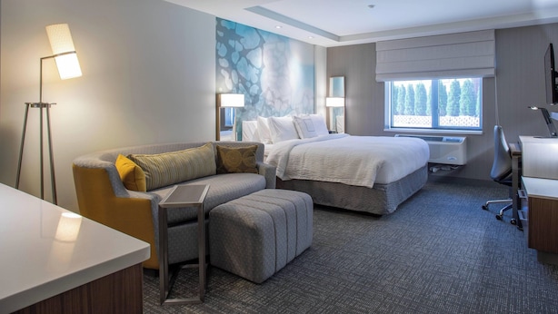A hotel room with a bed, a sofa, a desk, and a standing lamp. - Courtyard by Marriott New York Queens/Fresh Meadows (Fresh Meadows)
